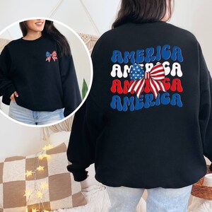 K&ouml;nnte beinhalten: Schwarzer Pullover mit einer rot-wei&szlig;-blauen Schleife und dem Wort "AMERICA", das viermal in einer Retro-Schrift wiederholt wird.