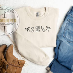 Peut inclure: Sweat-shirt cr&egrave;me brod&eacute; avec des dessins noirs : un n&oelig;ud, un c&oelig;ur, un ordinateur, un flacon pulv&eacute;risateur et un n&oelig;ud. Les mots "CADEAU PERSONNALIS&Eacute;" et "SWEAT-SHIRT & HOODIE BROD&Eacute;" sont dans un cercle. Bottes marron et jean bleu sont &eacute;galement dans l'image.