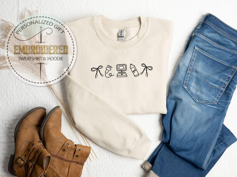 Peut inclure: Sweat-shirt cr&egrave;me brod&eacute; avec des dessins noirs : un n&oelig;ud, un c&oelig;ur, un ordinateur, un flacon pulv&eacute;risateur et un n&oelig;ud. Les mots "CADEAU PERSONNALIS&Eacute;" et "SWEAT-SHIRT & HOODIE BROD&Eacute;" sont dans un cercle. Bottes marron et jean bleu sont &eacute;galement dans l'image.