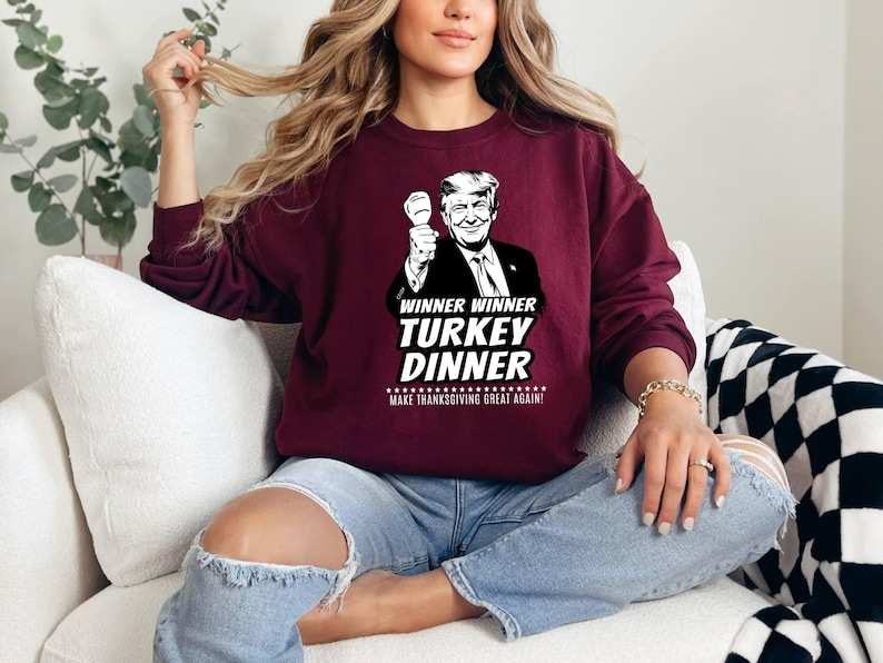 May include: Burgundy sweatshirt with a graphic of a man holding a fist up, with the text "WINNER WINNER TURKEY DINNER MAKE THANKSGIVING GREAT AGAIN!". The sweatshirt is a solid color and has long sleeves.
