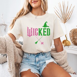 May include: Cream-colored t-shirt with the word "WICKED" in pink and green glitter lettering. The design includes a witch hat and crossed broomsticks. The shirt is paired with denim shorts.