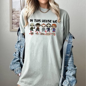 May include: A light green t-shirt with the text "In this house we" and a row of cartoon horror movie characters. The characters include Jason Voorhees, Freddy Krueger, Pennywise, Michael Myers, and Chucky. Each character has a speech bubble with text that says "Love People", "Where My Blood", "Like To Clown Around", "Love Family", and "Play With Dolls".
