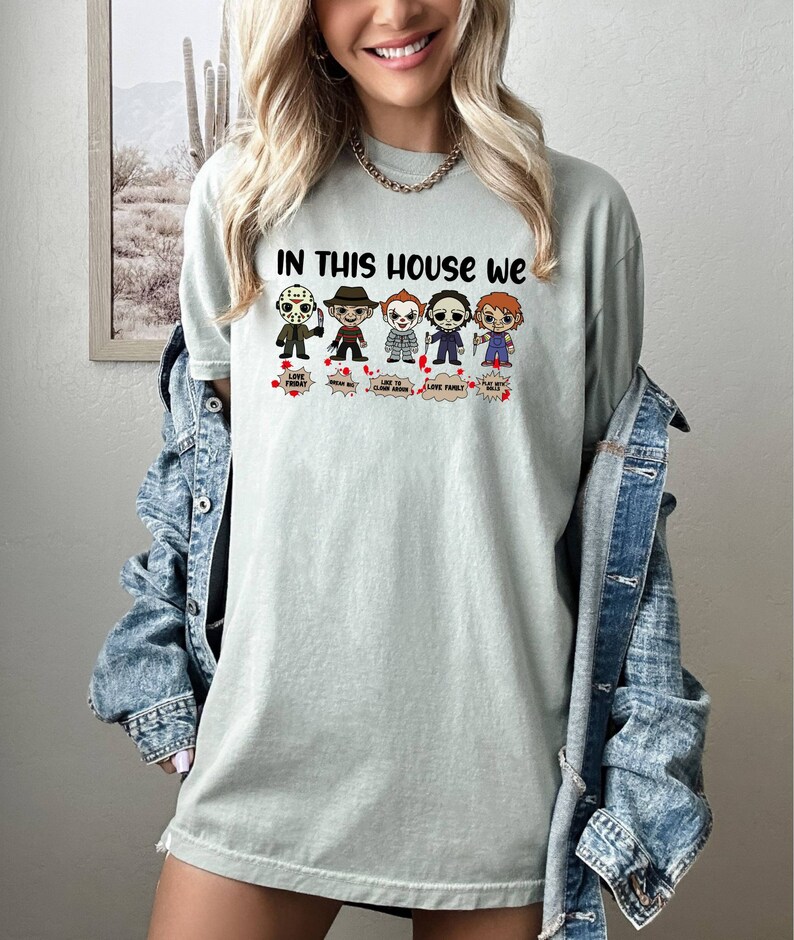 May include: A light green t-shirt with the text "In this house we" and a row of cartoon horror movie characters. The characters include Jason Voorhees, Freddy Krueger, Pennywise, Michael Myers, and Chucky. Each character has a speech bubble with text that says "Love People", "Where My Blood", "Like To Clown Around", "Love Family", and "Play With Dolls".