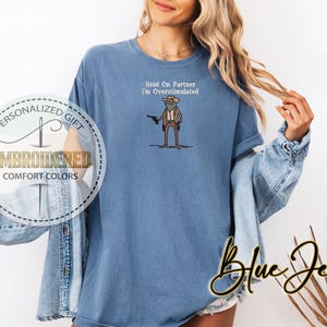 May include: A blue comfort colors t-shirt with the embroidered text "Hold On Partner I'm Overstimulated". The shirt features a cartoon frog wearing a cowboy hat and holding a gun. The shirt also has the text "Embroidered Comfort Colors" and "Personalized Gift" on the left side.