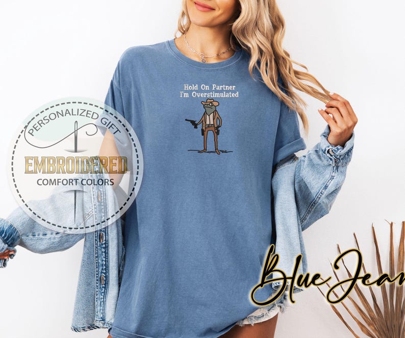 May include: A blue comfort colors t-shirt with the embroidered text "Hold On Partner I'm Overstimulated". The shirt features a cartoon frog wearing a cowboy hat and holding a gun. The shirt also has the text "Embroidered Comfort Colors" and "Personalized Gift" on the left side.
