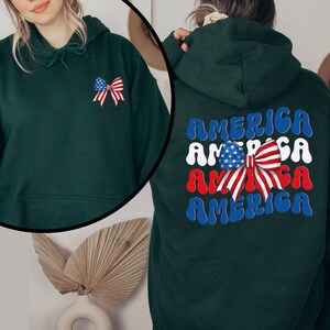 K&ouml;nnte beinhalten: Ein dunkelgr&uuml;ner Hoodie mit einer rot-wei&szlig;-blauen Schleife mit Sternen darauf. Auf der R&uuml;ckseite des Hoodies steht das Wort "AMERICA" viermal in einer wellenf&ouml;rmigen Schrift.