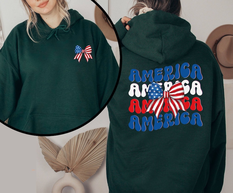 K&ouml;nnte beinhalten: Ein dunkelgr&uuml;ner Hoodie mit einer rot-wei&szlig;-blauen Schleife mit Sternen darauf. Auf der R&uuml;ckseite des Hoodies steht das Wort "AMERICA" viermal in einer wellenf&ouml;rmigen Schrift.