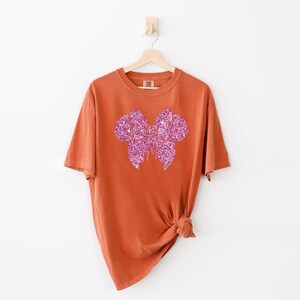 May include: A rust-colored t-shirt with a pink glitter bow design.