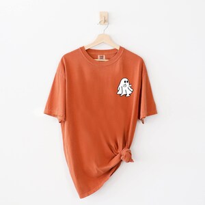 May include: A burnt orange t-shirt with a white ghost graphic on the front. The ghost is outlined in black and has a small smile.