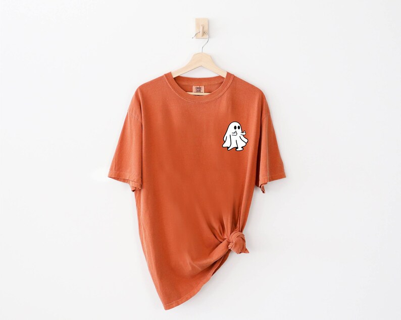 May include: A burnt orange t-shirt with a white ghost graphic on the front. The ghost is outlined in black and has a small smile.