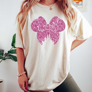 May include: A white t-shirt with a pink glitter bow design. The bow is in the shape of Mickey Mouse ears.