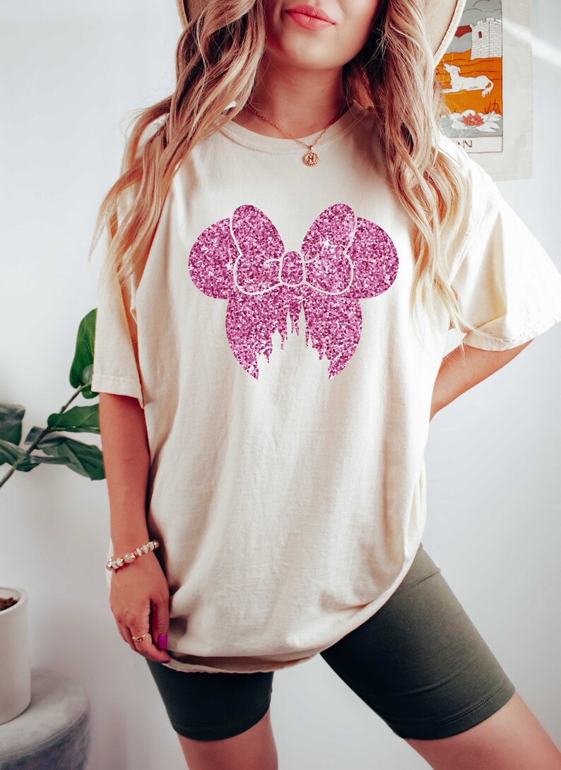 May include: A white t-shirt with a pink glitter bow design. The bow is in the shape of Mickey Mouse ears.