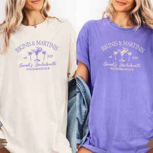 May include: Two t-shirts in ivory and violet with the text "Bikinis & Martinis est. 2024 Sarah's Bachelorette Tulum Beach Club" and a graphic of a cocktail glass with a lime and palm trees.