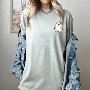 May include: A light green t-shirt with a white ghost graphic giving a thumbs up. The ghost has two black eyes and a small smile.