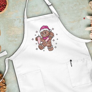 Peut inclure: Tablier de cuisine blanc avec un bonhomme en pain d'&eacute;pices portant un chapeau de P&egrave;re No&euml;l et tenant une boisson rose. Le bonhomme en pain d'&eacute;pices est entour&eacute; de petites &eacute;toiles blanches.