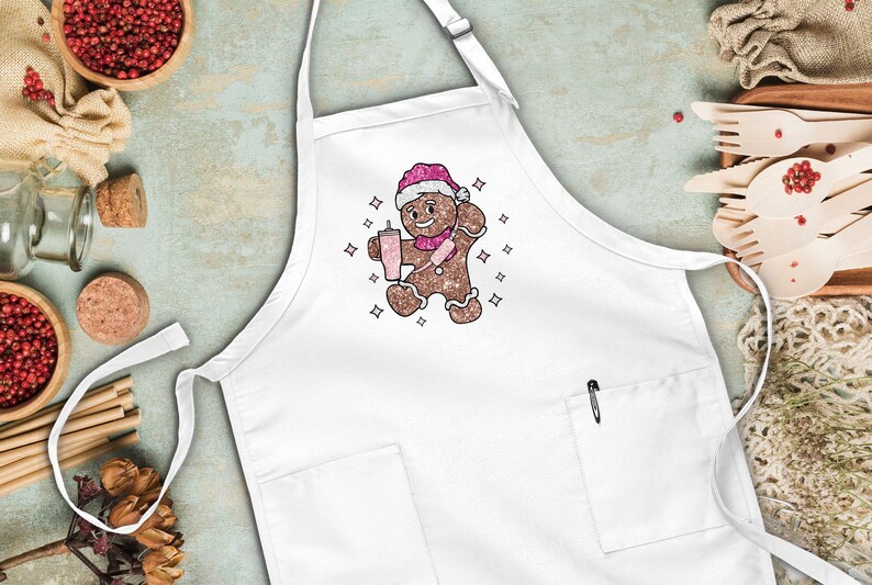 Peut inclure: Tablier de cuisine blanc avec un bonhomme en pain d'&eacute;pices portant un chapeau de P&egrave;re No&euml;l et tenant une boisson rose. Le bonhomme en pain d'&eacute;pices est entour&eacute; de petites &eacute;toiles blanches.