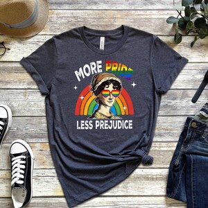 May include: A dark grey t-shirt featuring a rainbow and a portrait of a woman wearing rainbow heart-shaped sunglasses. The text "More Pride Less Prejudice" is printed on the shirt.