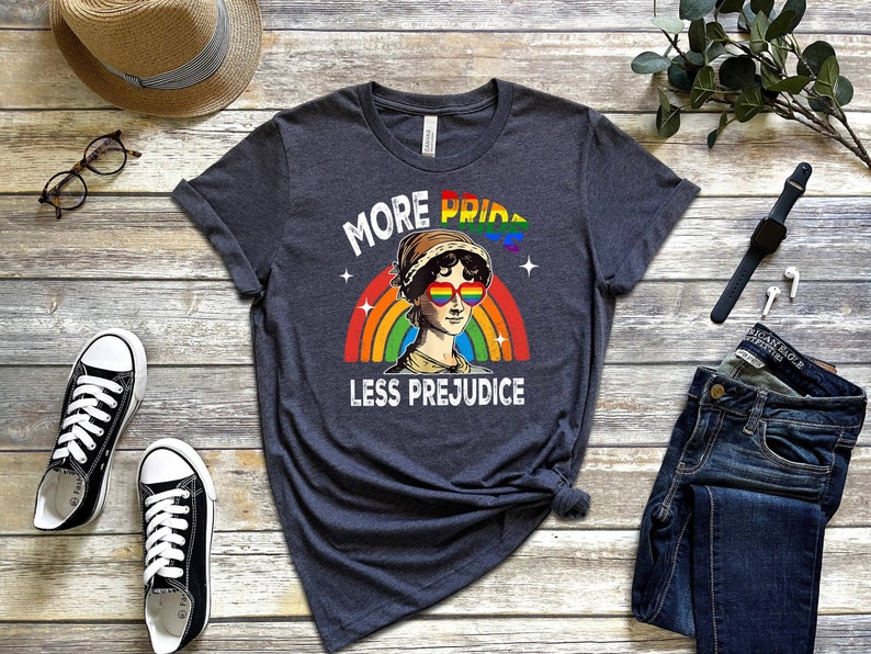 May include: A dark grey t-shirt featuring a rainbow and a portrait of a woman wearing rainbow heart-shaped sunglasses. The text "More Pride Less Prejudice" is printed on the shirt.