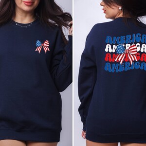 K&ouml;nnte beinhalten: Ein marineblauer Sweatshirt mit einer rot-wei&szlig;-blauen Schleife mit Sternen auf der Vorderseite und dem Wort "AMERICA" viermal in einer Retro-Schrift auf der R&uuml;ckseite.