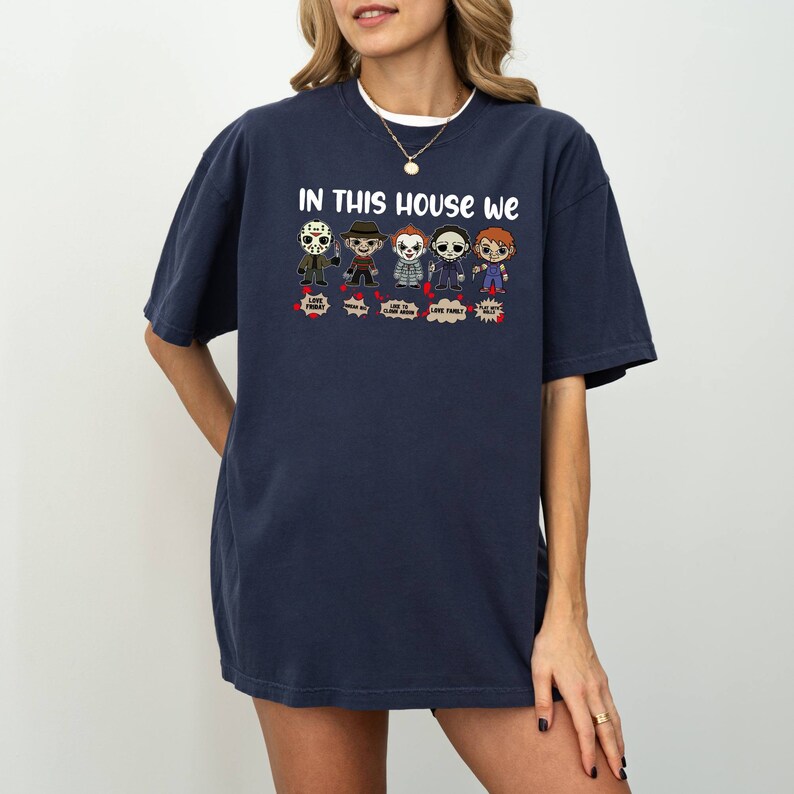 May include: A navy blue t-shirt with a graphic of horror movie characters. The text "In this house we" is printed above the graphic. The characters include Jason Voorhees, Freddy Krueger, Pennywise, Michael Myers, and Chucky.