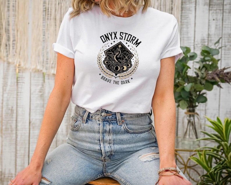 Puede incluir: Una camiseta blanca con un dise&ntilde;o gr&aacute;fico en negro y dorado que presenta un drag&oacute;n, lunas y estrellas. El gr&aacute;fico tiene el texto "ONYX STORM" y "BRAVE THE DARK".