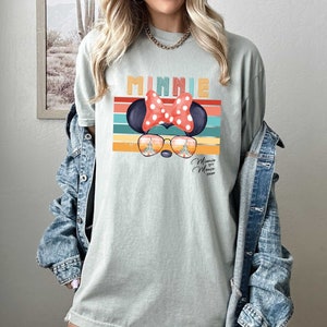 Op de afbeelding: Een lichtblauw T-shirt met een retro-grafische afbeelding van Minnie Mouse die een zonnebril draagt met een kasteelreflectie. De afbeelding bevat de tekst "Minnie Mouse 2023".
