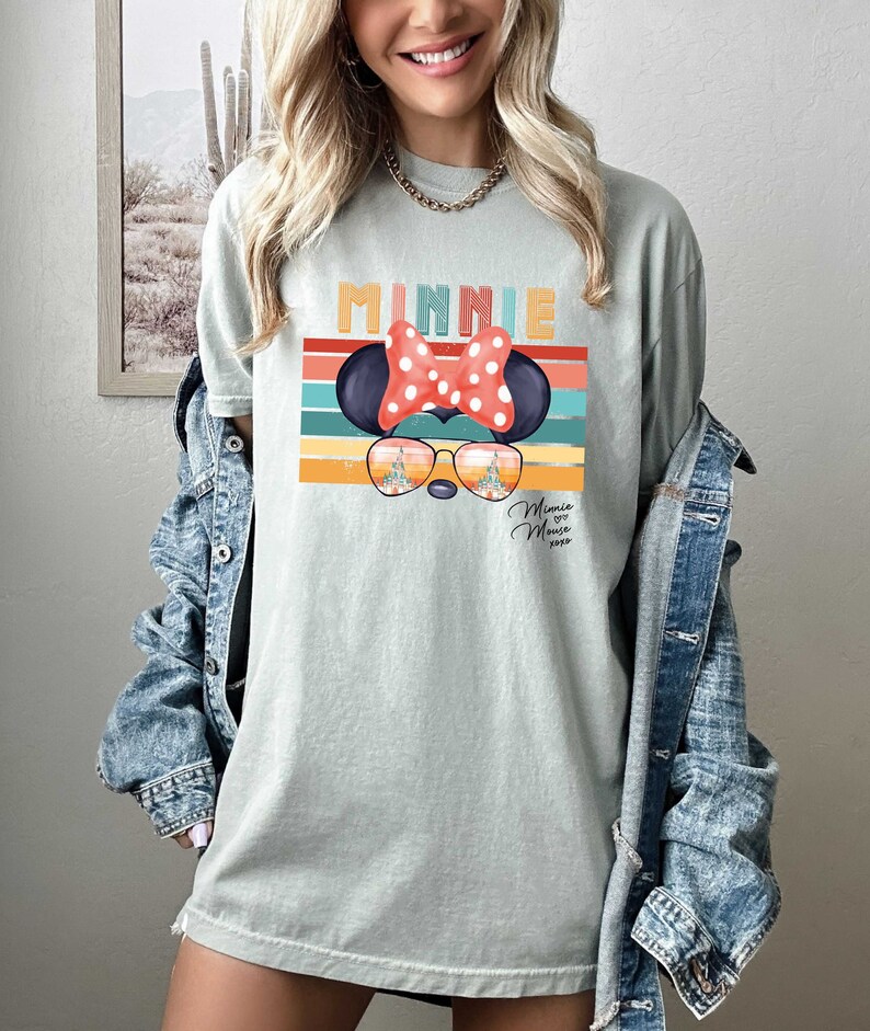 Op de afbeelding: Een lichtblauw T-shirt met een retro-grafische afbeelding van Minnie Mouse die een zonnebril draagt met een kasteelreflectie. De afbeelding bevat de tekst "Minnie Mouse 2023".