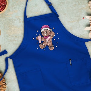 Peut inclure: Un tablier bleu avec un bonhomme en pain d'&eacute;pices tenant une boisson rose et portant un chapeau de P&egrave;re No&euml;l. Le bonhomme en pain d'&eacute;pices est entour&eacute; d'&eacute;toiles scintillantes blanches.