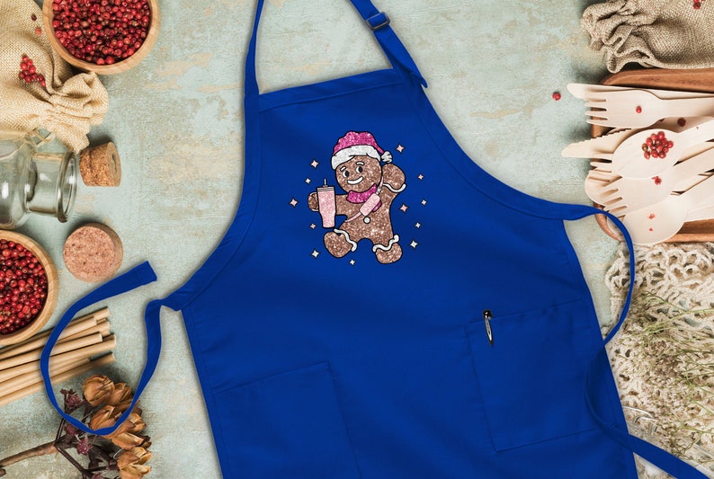 Peut inclure: Un tablier bleu avec un bonhomme en pain d'&eacute;pices tenant une boisson rose et portant un chapeau de P&egrave;re No&euml;l. Le bonhomme en pain d'&eacute;pices est entour&eacute; d'&eacute;toiles scintillantes blanches.