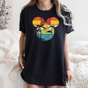 May include: Black t-shirt with a retro-style graphic of a Mickey Mouse head silhouette with palm trees, a sunset, and a beach chair. The graphic is in shades of yellow, orange, red, blue, and green.