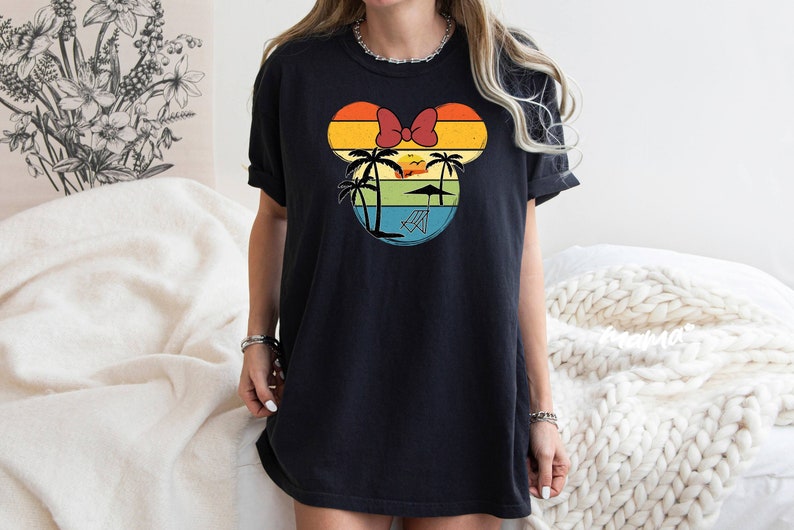 May include: Black t-shirt with a retro-style graphic of a Mickey Mouse head silhouette with palm trees, a sunset, and a beach chair. The graphic is in shades of yellow, orange, red, blue, and green.