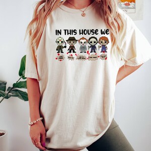 May include: A white t-shirt with the text "In this house we" and a row of cartoon horror movie characters. The characters include Jason Voorhees, Freddy Krueger, Pennywise, Michael Myers, and Chucky. Each character has a speech bubble with a phrase related to their character.