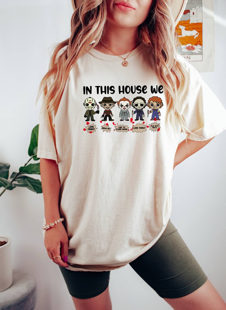 May include: A white t-shirt with the text "In this house we" and a row of cartoon horror movie characters. The characters include Jason Voorhees, Freddy Krueger, Pennywise, Michael Myers, and Chucky. Each character has a speech bubble with a phrase related to their character.