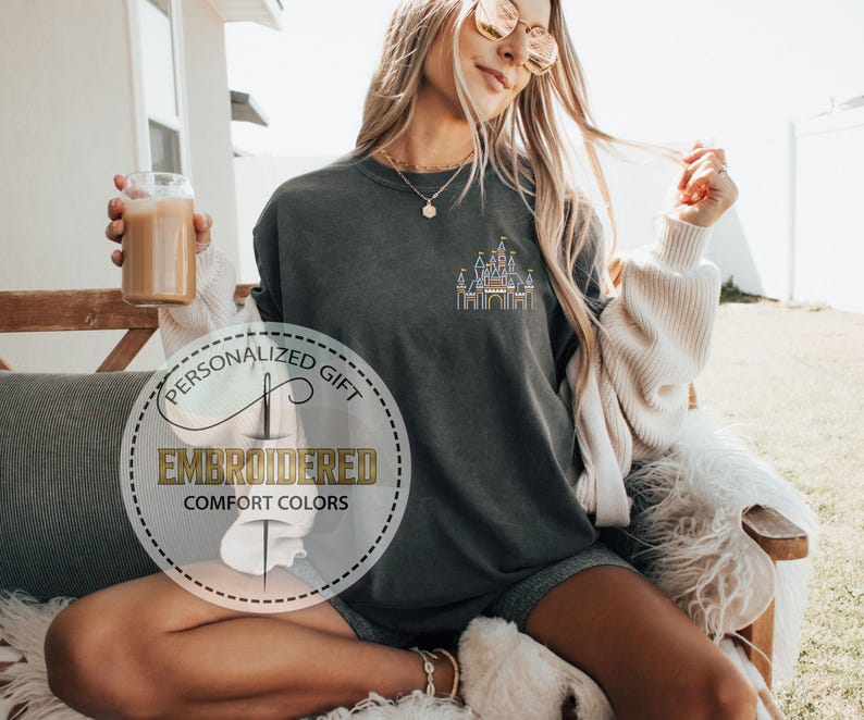 May include: A dark gray t-shirt with a white embroidered design of a castle. The t-shirt has the text "Personalized Gift" and "Embroidered Comfort Colors" on a white circle.