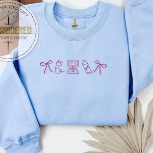 Peut inclure: Sweat-shirt bleu clair avec broderie rose. Le motif comprend un n&oelig;ud, un c&oelig;ur avec des ondes sonores, un ordinateur, une bouteille avec le mot "gel" et un autre n&oelig;ud. Le sweat-shirt porte les inscriptions "PERSONALIZED GIFT" et "EMBROIDERED SWEATSHIRT & HOODIE".
