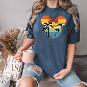 May include: A navy blue t-shirt with a retro-style graphic of a cartoon mouse head with a red bow. The graphic features palm trees, a sunset, and a beach chair. The text "The Fast Pool" is visible in the background.