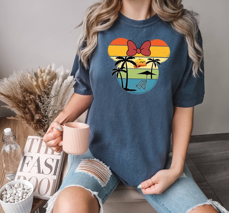 May include: A navy blue t-shirt with a retro-style graphic of a cartoon mouse head with a red bow. The graphic features palm trees, a sunset, and a beach chair. The text "The Fast Pool" is visible in the background.