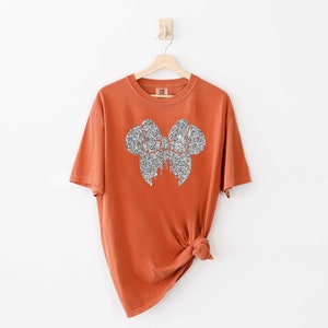 May include: A burnt orange t-shirt with a silver glitter bow design.