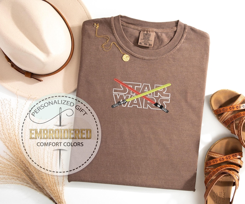 May include: A brown t-shirt with the words "STAR WARS" embroidered on the front. The words are in white and the embroidery includes two crossed lightsabers, one red and one yellow.