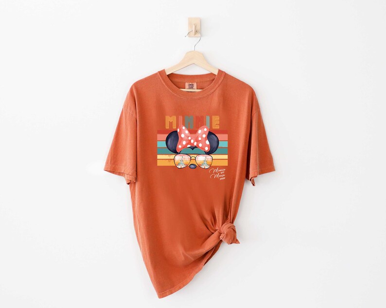 Op de afbeelding: Een oranje T-shirt met een retro-stijl afbeelding van Minnie Mouse met een zonnebril en een rode strik. De afbeelding bevat de tekst "Minnie Mouse" en "2023".