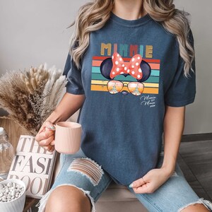 Op de afbeelding: Een marineblauw T-shirt met een retro-grafische afbeelding van Minnie Mouse met een zonnebril en een rode strik. De afbeelding bevat de tekst "Minnie Mouse 2023".