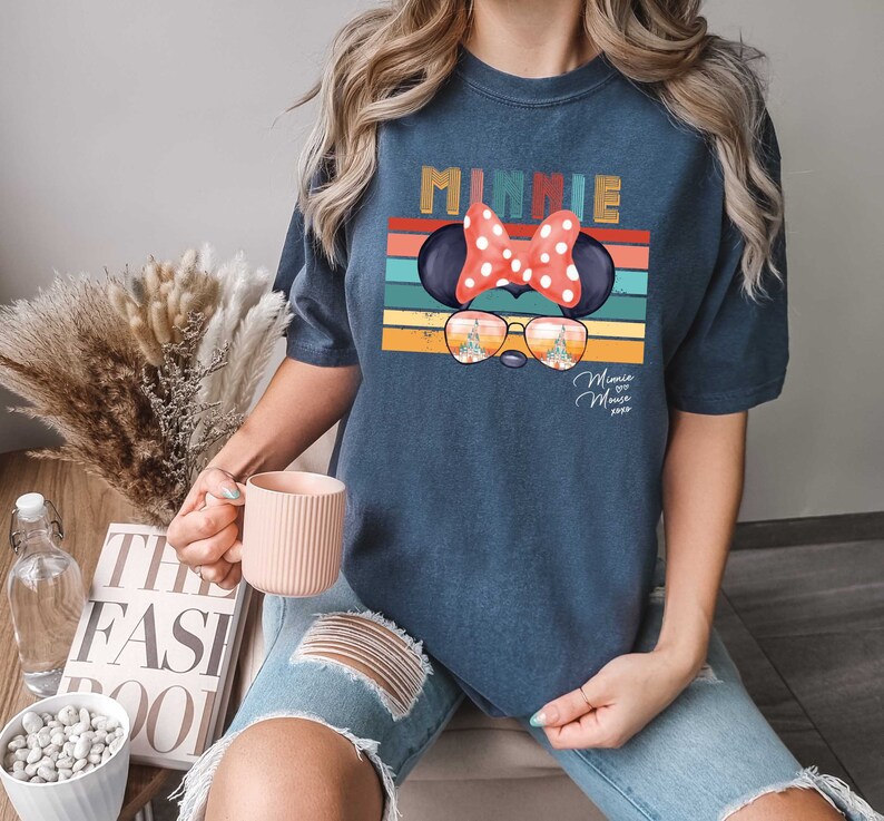 Op de afbeelding: Een marineblauw T-shirt met een retro-grafische afbeelding van Minnie Mouse met een zonnebril en een rode strik. De afbeelding bevat de tekst "Minnie Mouse 2023".
