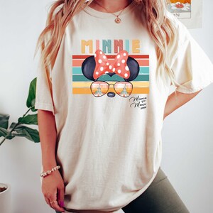 Op de afbeelding: Een wit T-shirt met een retro-stijl grafische afbeelding van Minnie Mouse met een zonnebril. De afbeelding bevat de tekst "Minnie" en "Minnie Mouse xoxo".