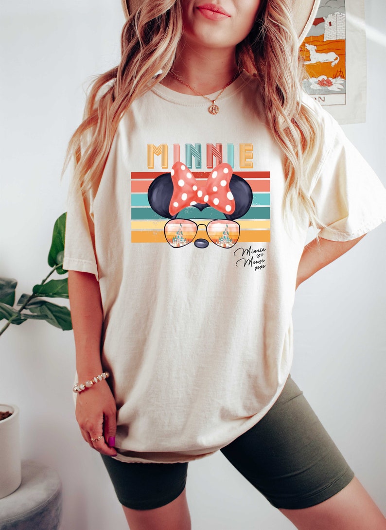 Op de afbeelding: Een wit T-shirt met een retro-stijl grafische afbeelding van Minnie Mouse met een zonnebril. De afbeelding bevat de tekst "Minnie" en "Minnie Mouse xoxo".