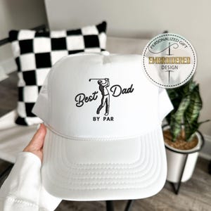May include: White trucker hat with a mesh back and a curved brim. The hat features the text "Best Dad by Par" in black script, along with a graphic of a golfer. A circular emblem reads "Personalized Gift, Embroidered Design."