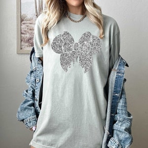 May include: A light green t-shirt with a silver glitter bow design.