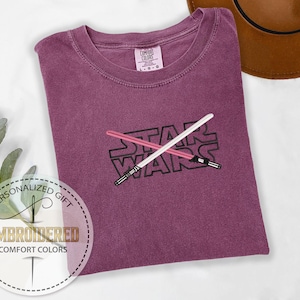 May include: A purple t-shirt with the words "STAR WARS" embroidered on the front. Two lightsabers are crossed behind the text.