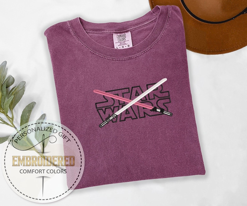 May include: A purple t-shirt with the words "STAR WARS" embroidered on the front. Two lightsabers are crossed behind the text.