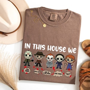May include: A brown t-shirt with the text "In this house we" and a row of cartoon horror movie characters. The characters include Jason Voorhees, Freddy Krueger, Pennywise, Michael Myers, and Chucky. Each character has a speech bubble with a phrase related to their movie.