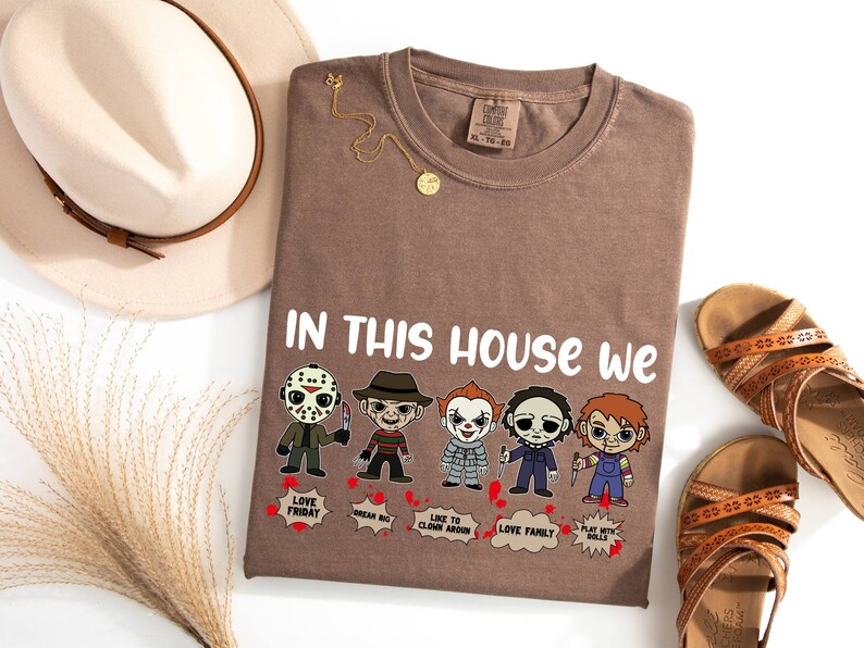 May include: A brown t-shirt with the text "In this house we" and a row of cartoon horror movie characters. The characters include Jason Voorhees, Freddy Krueger, Pennywise, Michael Myers, and Chucky. Each character has a speech bubble with a phrase related to their movie.